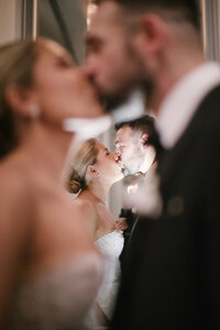 Bride and groom kiss, their reflection captured softly in a mirrored surface