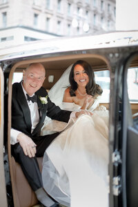Bride laughs and waves from inside a vintage car seated beside her father