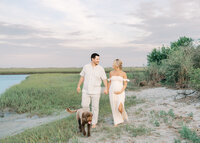 A couple walking along the beach with their dog in the backdrop during their maternity photography session at Wrightsville Beach, North Carolina