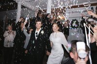 Laughing bride and groom exit through a glittering cascade of bubbles surrounded by cheering guests at night