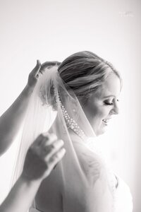 A bride smiles gently as someone adjusts her veil. The image is in black and white, capturing a soft, intimate moment before the wedding ceremony.