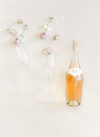 Elegant wedding reception detail featuring a bottle of rosé surrounded by delicate blush and ivory flowers in minimalist glass bud vases.