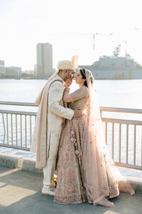 South Asian bride and groom in ornate traditional attire share an intimate forehead-touch beside the Norfolk harbor at golden hour