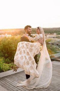 Groom lifts laughing bride in gold-embroidered lehenga on a rooftop terrace bathed in warm sunset light