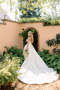 Smiling bride in a sweeping ivory ballgown glances over her shoulder before a vine-draped brick archway in a lush garden