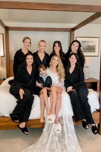 Smiling bride in lace robe and fluffy slippers sits with six bridesmaids in black pajamas on a four-poster bed