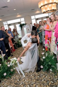 Groom dramatically dips and kisses his bride in the flower-lined aisle as cheering guests celebrate at Belmont Country Club
