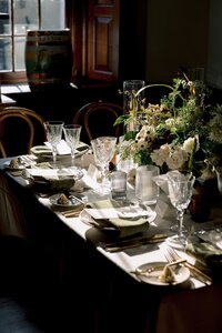 Moody sunlit wedding dinner table set with crystal glassware, gold cutlery, sage napkins, and lush floral centerpiece in a dark elegant venue