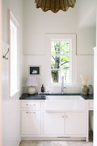 Bright white farmhouse kitchen with apron-front sink beneath a sunny window, shaker cabinets, plants, and warm vintage decor creating a calm mood.