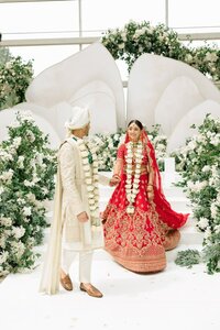 Bride in red gold lehenga and groom in ivory sherwani exchange garlands at a lush white floral mandap indoors