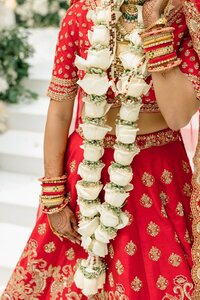 Indian bride in red gold embroidered lehenga holds white rose varmala garland adorned with bangles and mehndi during ceremony