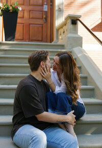 Engaged couple sitting on steps in Boston's Back Bay with bride holding grooms face and smiling.