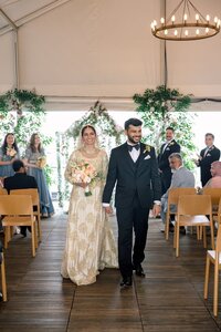 Radiant South Asian bride in gold-embroidered lehenga and groom in black tuxedo walk hand-in-hand down a flower-adorned aisle