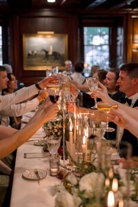 Wedding guests raise crystal glasses in a joyful toast along a candlelit dinner table inside a moody wood-paneled dining room