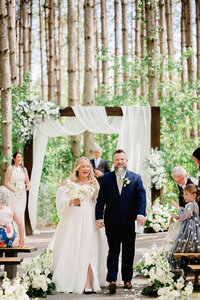 Bride and groom holding hands at the end of a wedding ceremony in the forest.