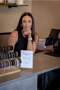 Woman jewelry designer smiles behind a display of colorful bracelets, with a handwritten sign and podcast microphone on the studio table.
