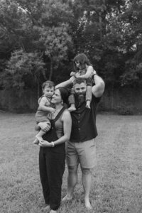 Father holds toddler on shoulders while mother holds toddler close to her face in their backyard during their at home family photo session near houston texas