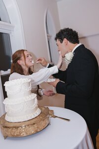 Laughing bride and groom smear wedding cake on each other beside a tiered white cake at their intimate chapel reception