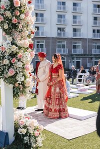 Bride in an ornate red lehenga and groom in a cream sherwani and red turban standing together at their outdoor ceremony beside a floral mandap, guest chairs and the venue behind them, at a Hyatt Chesapeake Bay Sikh wedding.
