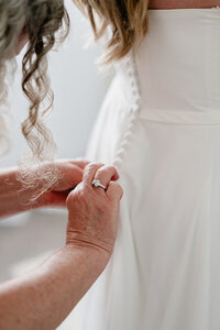 Mother of the bride helping a bride do up her buttons on her wedding dress while taking photos.