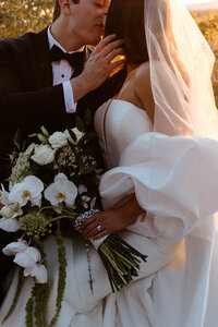 Close-up of bride and groom at golden hour with white orchids, roses, greenery and the bride's veil illuminated by sunlight.