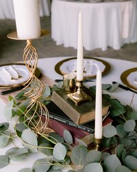 Table set with gold chargers and white plates and a centerpiece of gold candlesticks with white candles, a tall stack of vintage books and a halo of greenery