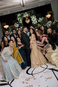 South Asian bride in gold lehenga laughs with groom and jubilant guests on monogrammed ballroom dance floor