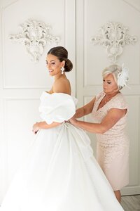 A smiling bride in an off-shoulder white gown stands as an older woman in a light pink dress helps adjust her dress. They are in a bright, elegant room with white ornate decor on the wall.