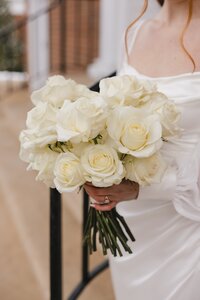 A bride in a white satin gown holds a lush ivory rose bouquet outside Liberty University Chapel in a timeless, elegant moment