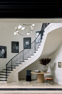 Organic, modern foyer with sweeping white staircase, black railings, sculptural chandelier, framed art, round wood table, and dried flowers.