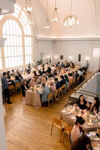 Wedding guests dining together at candlelit tables inside a grand DC venue with soaring arched windows and warm golden light