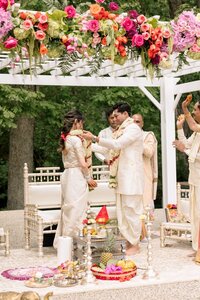 Bride and groom exchange floral garlands beneath a vibrant bloom-draped white mandap during an outdoor Hindu wedding ceremony
