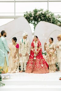 Smiling Indian bride in red lehenga and groom in ivory sherwani exchange garlands beneath a white floral archway at an elegant indoor ceremony