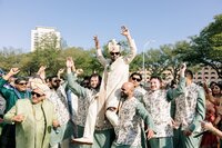 Jubilant Indian groom in ivory sherwani and turban raised high by groomsmen during a lively outdoor baraat procession under clear blue skies