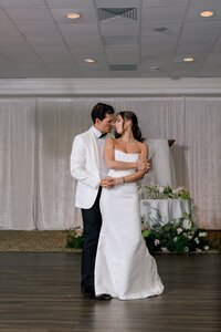 Bride and groom share a tender first dance on the hardwood floor of an elegant ballroom adorned with lush florals
