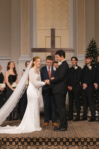 Laughing bride and groom exchange rings before a wooden cross at an intimate chapel wedding ceremony