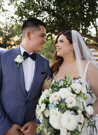 Elegant bridal portrait featuring a joyful couple sharing a quiet moment beneath oak trees with lush white florals, showcasing natural emotion and fine art wedding photography in San Antonio.