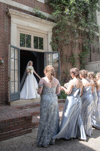 Bride stands in an open doorway framed by wisteria as bridesmaids look on in blue gowns