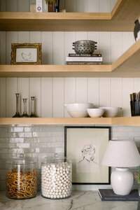 Warm, curated kitchen shelves display framed sketches, vintage books, metal bowl, white ceramics, pasta jars, beads, and a lamp.