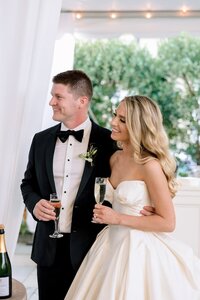 Smiling bride in strapless gown and tuxedoed groom holding champagne flutes inside an airy white tent with lush greenery beyond