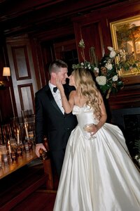 Bride and groom lean in for a kiss holding champagne and whiskey inside a dark wood-paneled Virginia manor