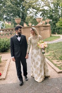 Smiling Muslim bride in gold-embroidered gown and groom in tuxedo walk hand-in-hand along a formal garden terrace in Washington DC