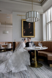 Bride and groom sit together in a restaurant booth beneath a gold framed portrait painting