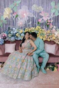South Asian bride and groom share a tender kiss on a velvet sofa surrounded by lush pastel blooms at their sangeet celebration