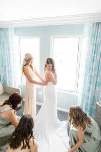 Bride getting ready with her bridesmaids as they fasten her wedding gown and arrange the train in a light-filled room.