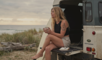 Lauren sitting on the beach with a surfboard, holding a cup of coffee