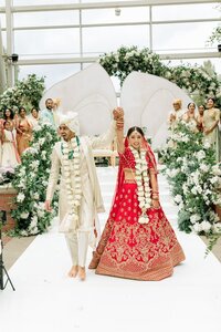 Beaming South Asian newlyweds raise joined hands while recessing down a white aisle framed by lush floral archways at an indoor garden ceremony