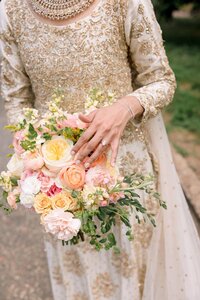 Muslim bride in gold embroidered gown cradling a lush peach and pink rose bouquet with diamond engagement ring visible