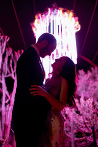 Newlywed couple embracing beneath a dramatic pink illuminated installation at their wedding reception.