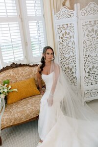 Elegant bride in strapless gown and flowing veil seated on an antique settee in a sunlit, ornate bridal suite